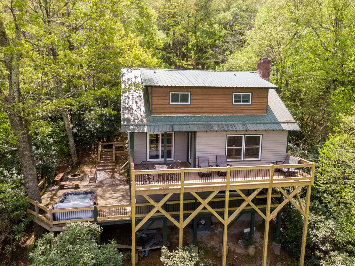 The hero image shows the rustic exterior of a cozy cabin nestled among lush green trees, featuring a spacious wooden deck with a bubbling hot tub. The deck offers comfortable seating and sweeping views of the serene natural surroundings, inviting guests to unwind and soak in mountain tranquility.