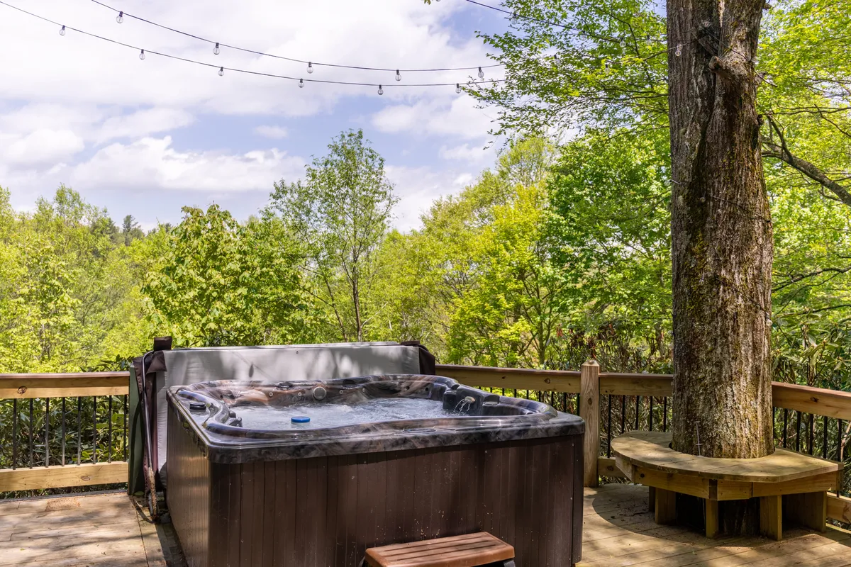 A tranquil wooden deck scene with a hot tub as the focal point, surrounded by lush greenery and illuminated by string lights. The setting evokes a peaceful retreat where guests can enjoy morning coffee, evening soaks, or quiet moments in fresh mountain air.
