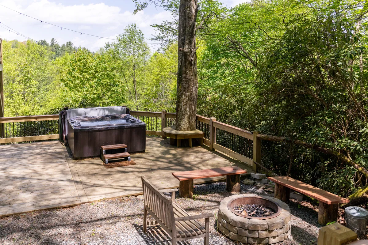 This image captures a serene outdoor setting featuring a bubbling hot tub and a nearby fire pit, surrounded by lush greenery and wooden benches. The space is designed for relaxing, social gatherings, and enjoying nature with a view of the deck area and expansive mountain surroundings.