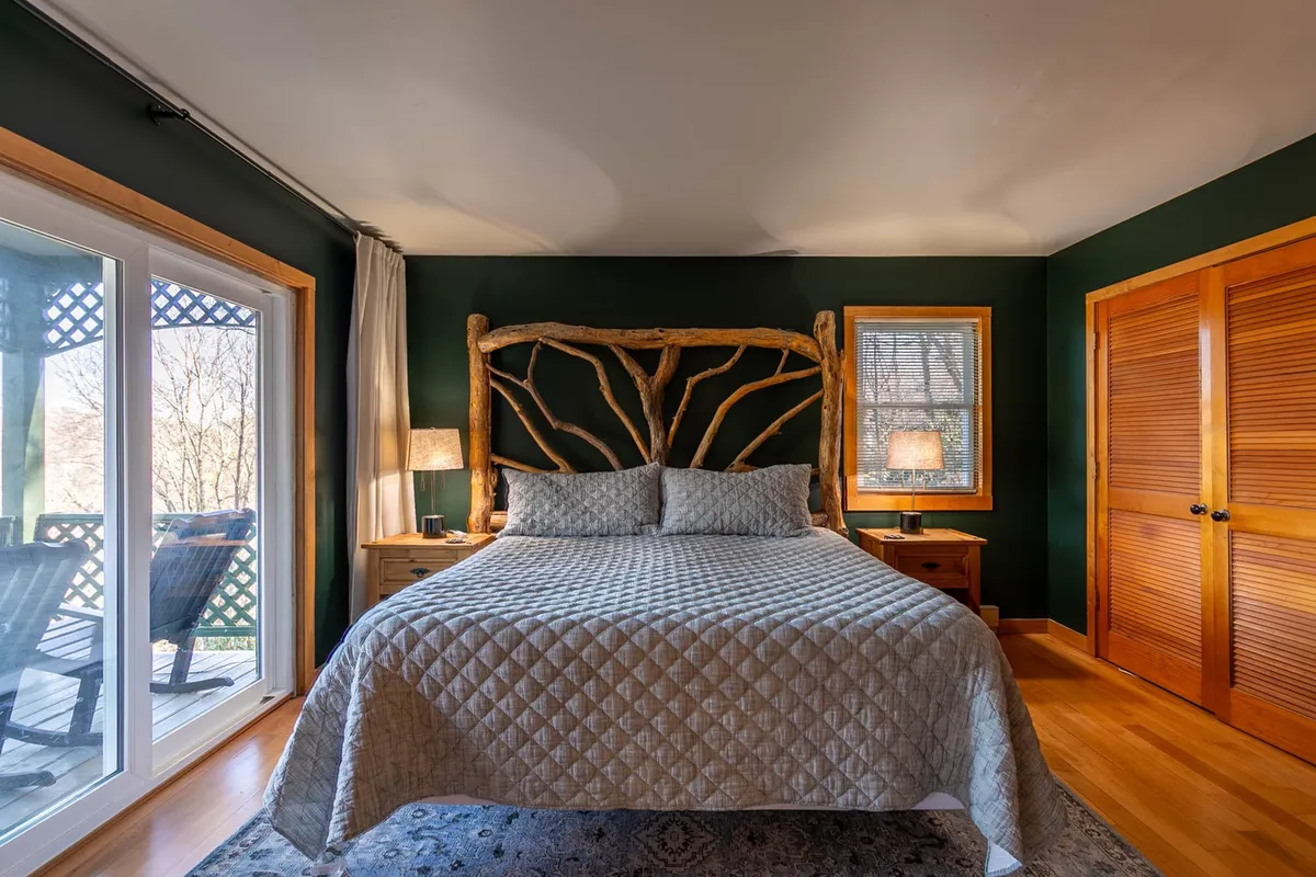 A tranquil bedroom featuring a wooden headboard, plush bedding, large windows, and wooden furniture. Soft morning light filters in, and the room offers calming views of the surrounding woodlands.