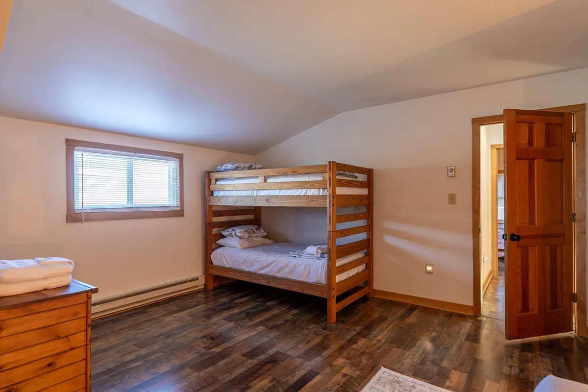 A cozy upstairs bedroom with twin bunk beds, a wooden dresser, and a window with blinds. The space feels playful yet restful, perfect for families or groups of children to unwind and build lasting vacation memories.