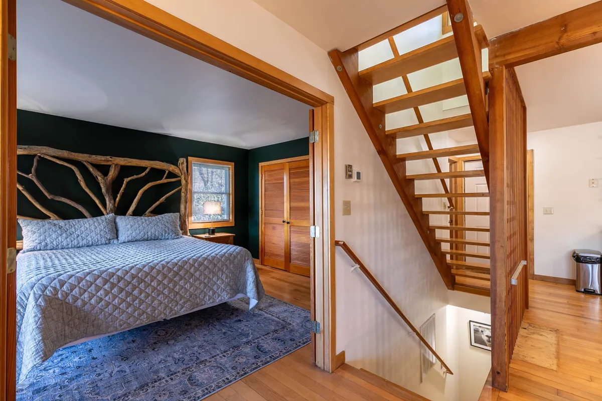 This image shows a serene bedroom featuring a bed with a quilted cover, a unique branch headboard, and a staircase leading up. The room is bathed in natural light, creating a tranquil atmosphere ideal for relaxation, reading, and restful sleep.