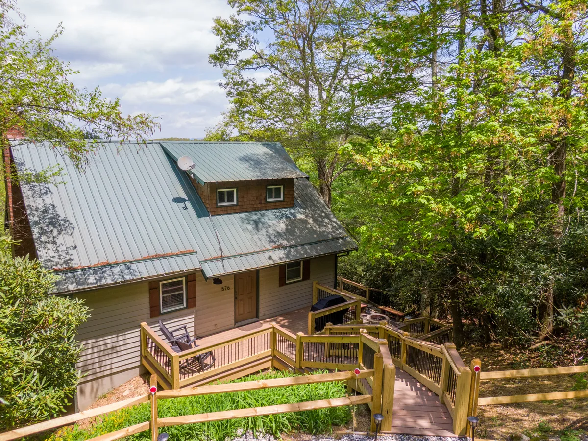 The wide wooden deck and metal roof of the home are featured, framed by lush green foliage and the inviting front door. The space suggests a perfect spot for family gatherings, nature walks, and outdoor meals, reinforcing the outdoorsy, adventure-driven theme of the section.