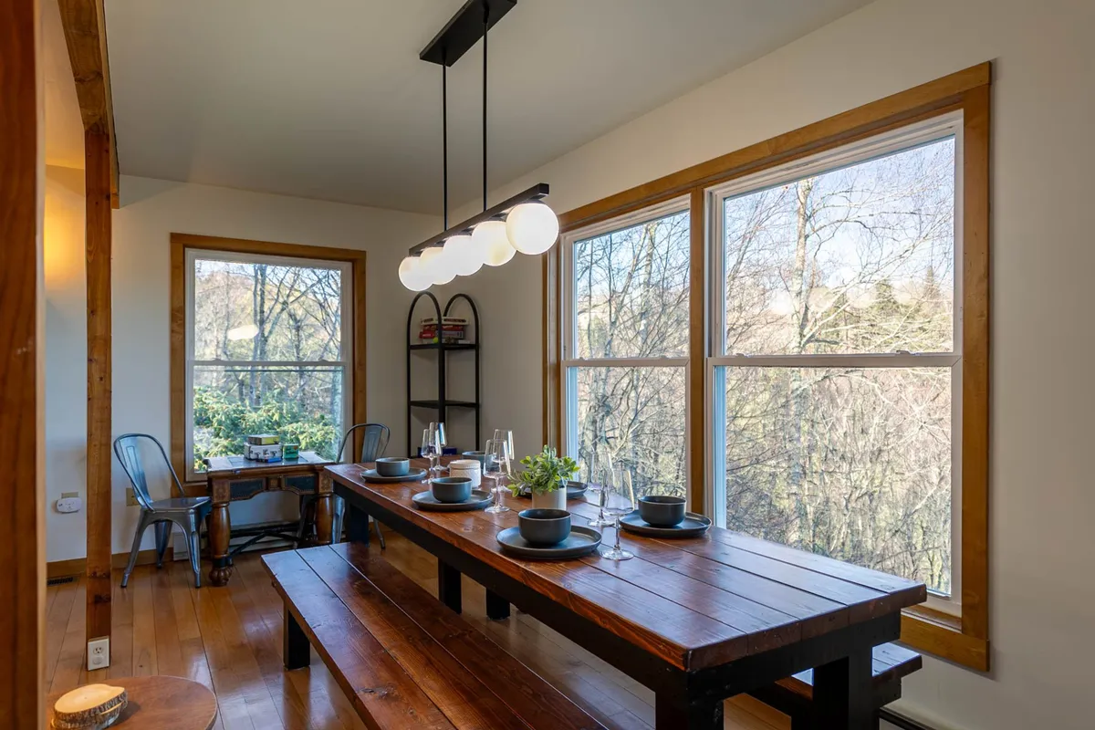 A long wooden dining table sits beneath a warm hanging light fixture, set by expansive windows that frame beautiful natural views. The scene is inviting and clearly designed for family dinners and celebrations, with enough seating for larger groups.