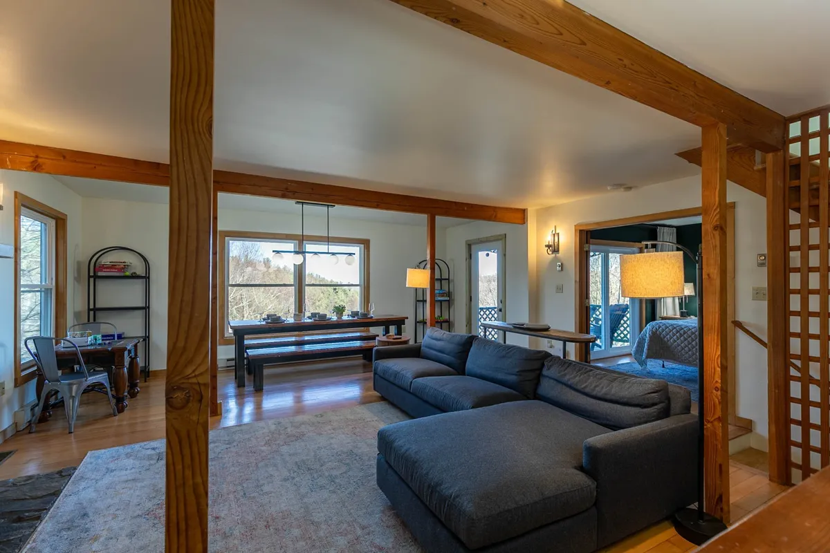 This image features a large sectional sofa and a dining table positioned near expansive windows that frame stunning views of the outdoors. The cozy seating area invites relaxation while the windows connect directly to the landscape beyond, hinting at the mountain views and the tranquil setting described in the section.