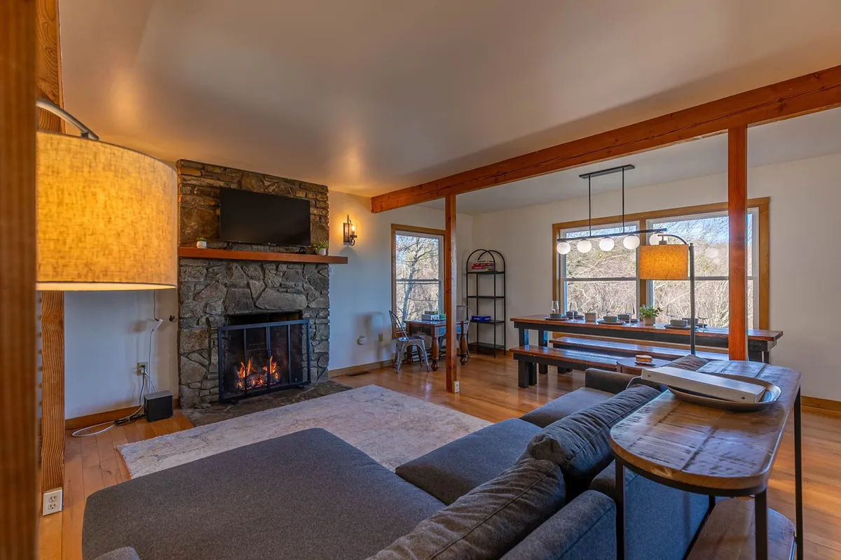 This image showcases a cozy living area with a fireplace, large window revealing mountain views, a plush sofa, and a spacious dining table. The abundant natural light and open layout highlight both the living and dining spaces, perfect for gatherings and relaxation.