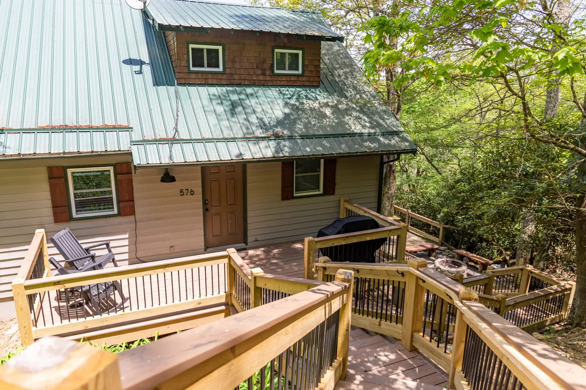 The welcoming front deck features steps rising to the green-roofed house surrounded by trees. This entryway signals the start of a memorable group adventure and hints at amenities like the fire pit and nature walks mentioned in the section.