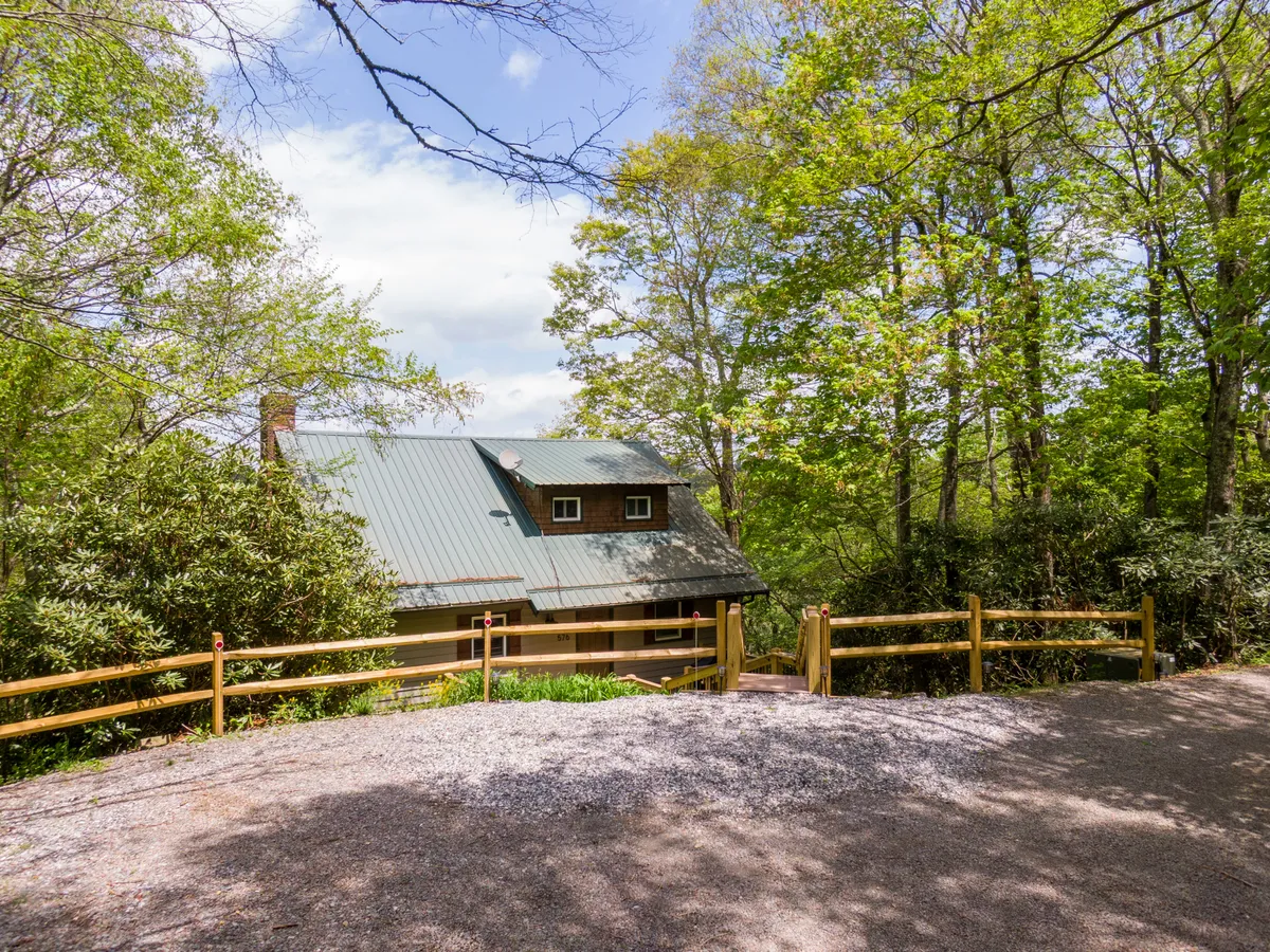 This exterior image showcases the house surrounded by trees and a rustic fence, giving a sense of seclusion and immersion in nature. The porch is visible, inviting relaxation and offering a clear connection between the home and the great outdoors.